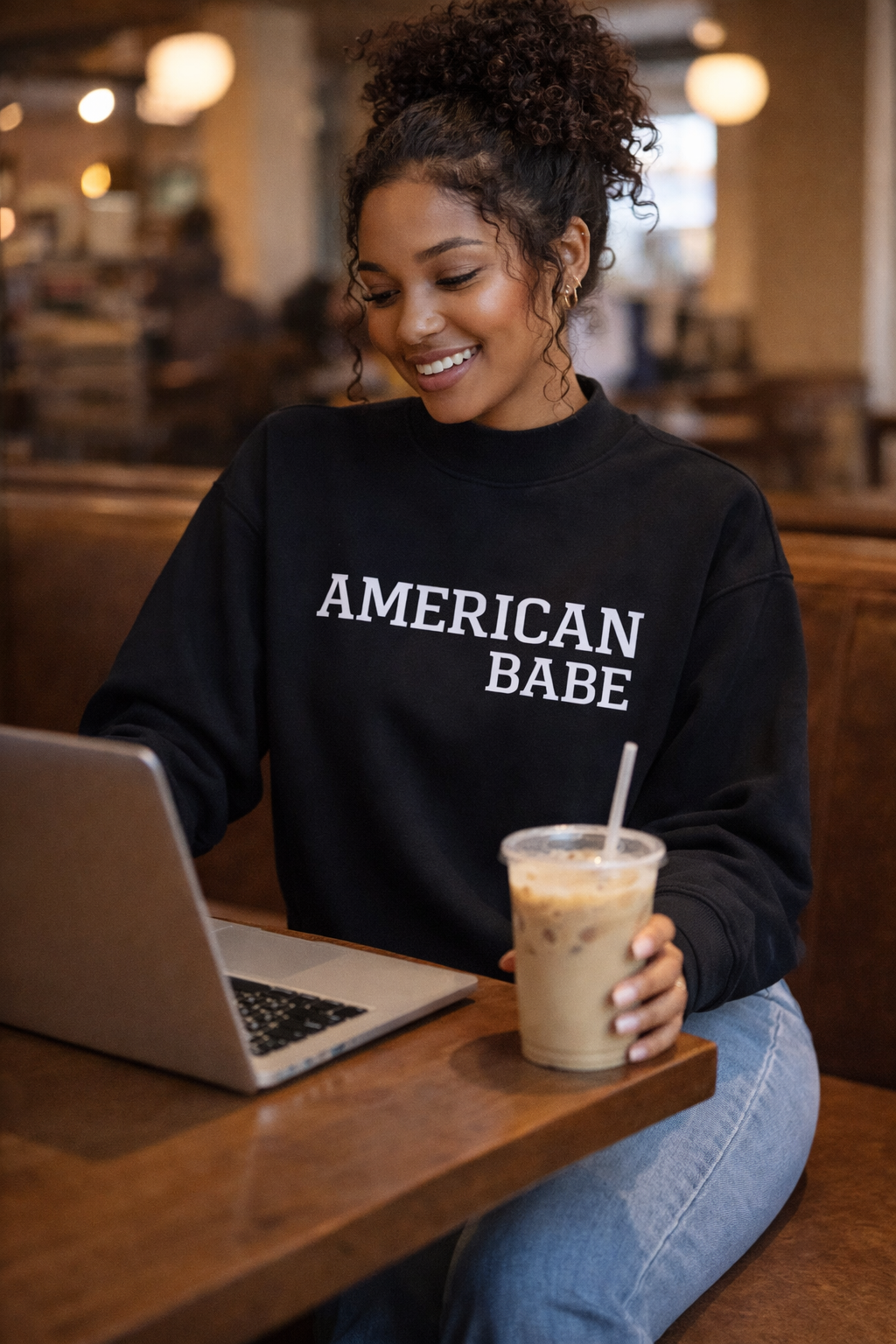 Woman wearing a black sweatshirt with 'American Babe' text, sitting at a table with a laptop and iced coffee.
